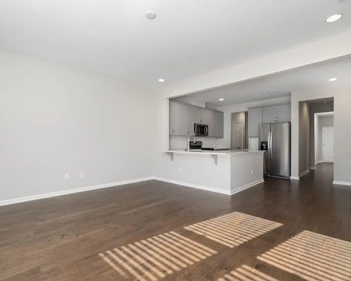 Open-plan room finished in white beside a kitchen, with sunlight across a dark timber floor